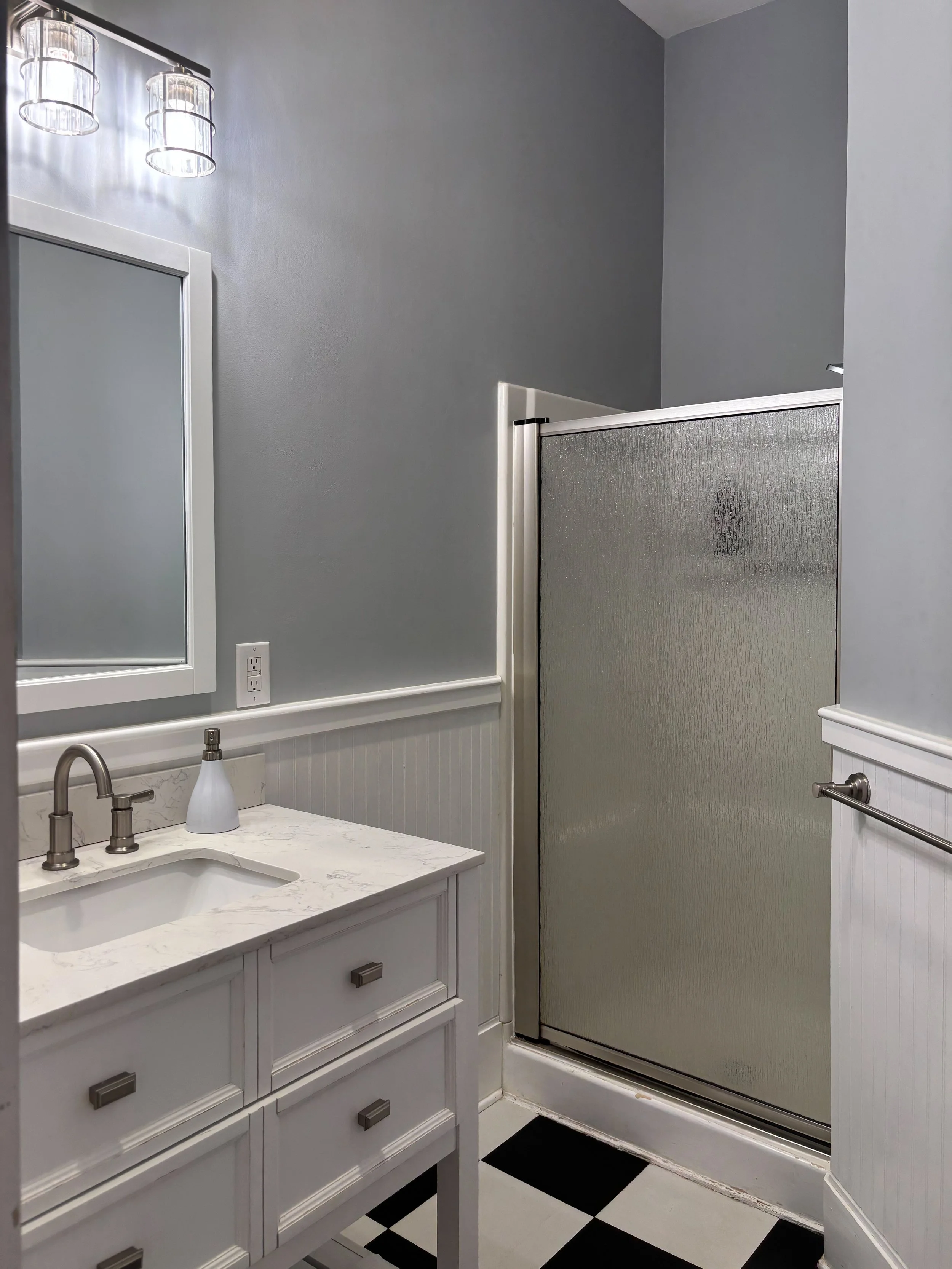 A small bathroom with a white vanity and sink, a mirror, gray walls, a frosted shower partition, and checkered black and white floor tiles.