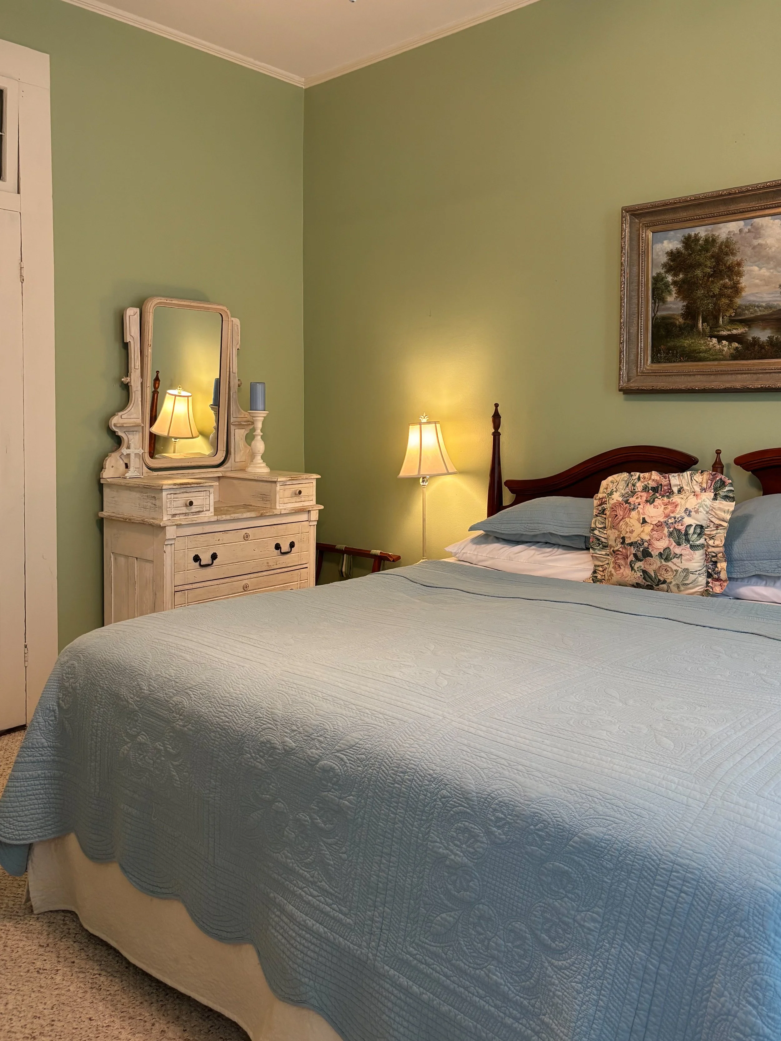 A bedroom with a bed covered in a light blue quilt, a floral pillow, and a nightstand with a mirror, lamp, and a candleholder, against a green wall with a framed landscape painting.