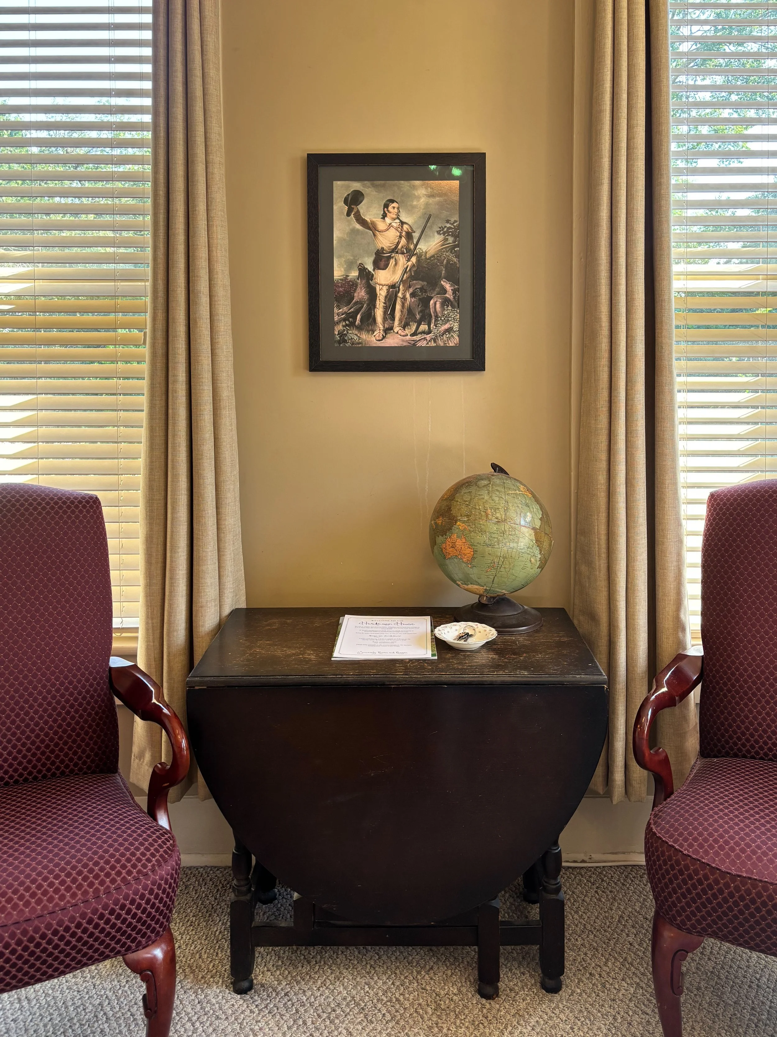A small table with a globe, a menu, and a small dish with keys, flanked by two upholstered armchairs, with a framed historic illustration on the wall and two windows with blinds and curtains.