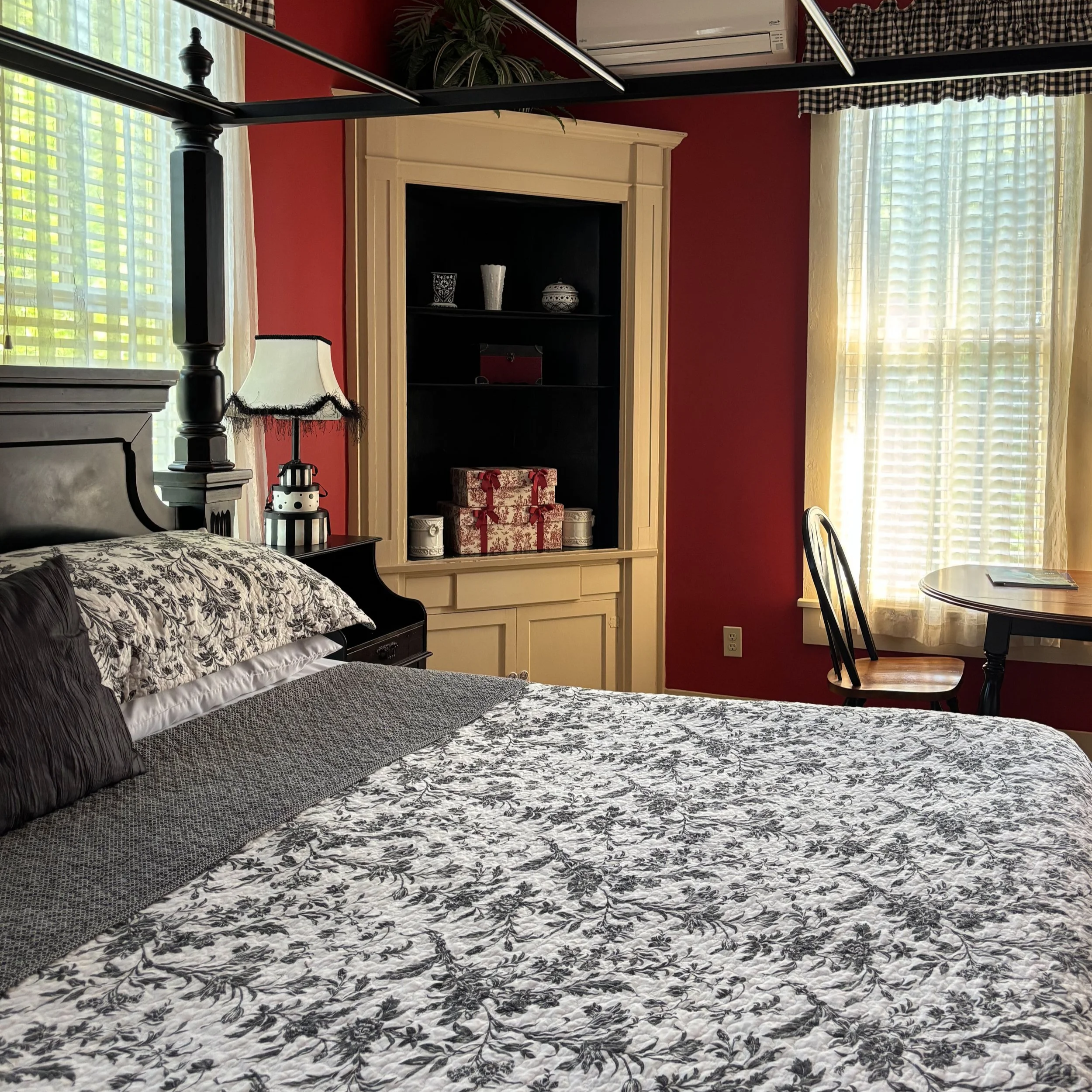 A bedroom with a bed covered in black and white floral bedding, a black and white dresser, a white fireplace mantel, a black bookcase with decorative items and Christmas presents, windows with white and checkered curtains, and a dining table with chairs.
