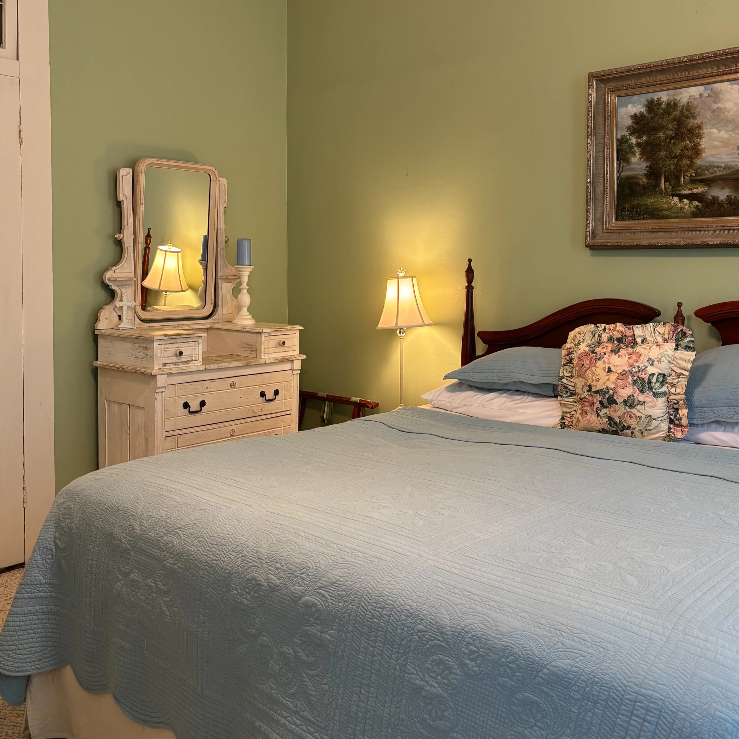 A bedroom with light green walls, a wooden headboard, a floral pillow, and a quilted bedspread. There is a white dresser with a mirror, a lamp, and a candle holder with a tall candle reflected in the mirror. A framed landscape painting of trees and a river hangs on the wall.