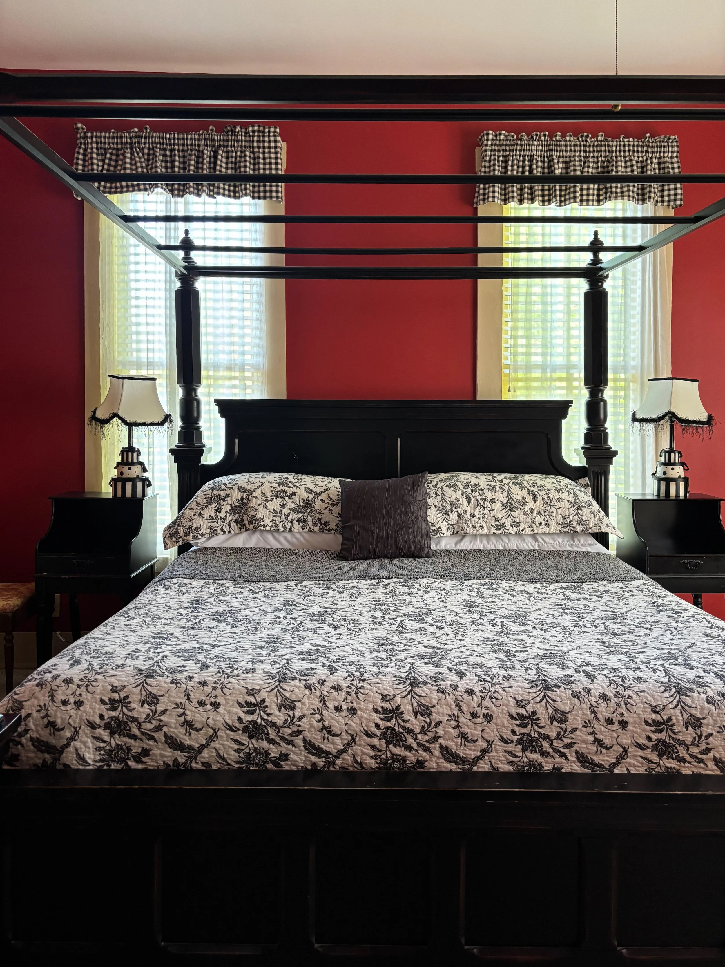 A bedroom with a black four-poster bed, floral bedspread, and checkered window valances. Two nightstands with lamps flank the bed, with two large windows behind with sheer curtains and yellow drapes.