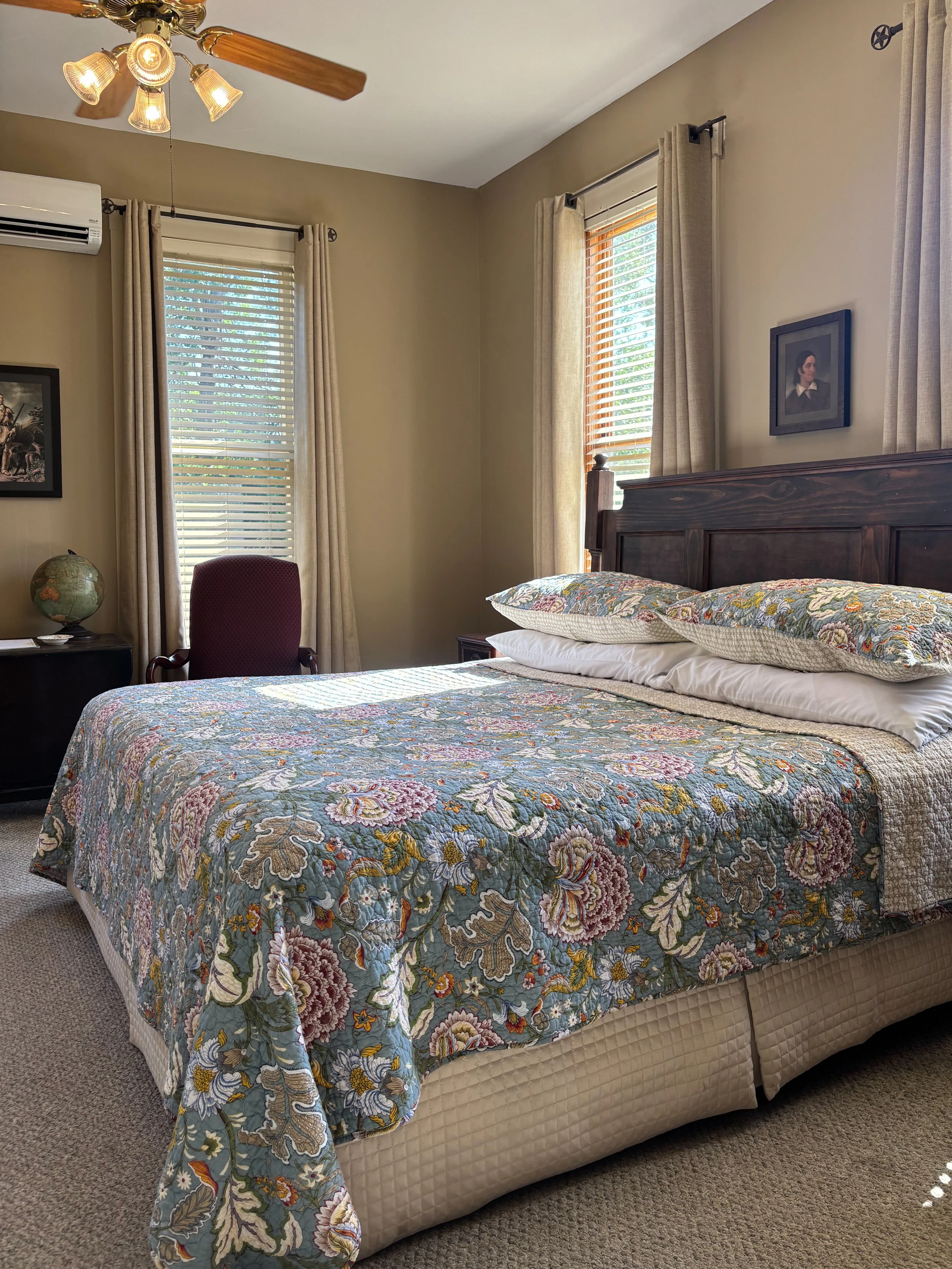 A bedroom with a bed covered in a floral quilt, a dark wooden headboard, and two pillows. There is a window with blinds and curtains on each side, a ceiling fan with lights, and a small side table with a globe and a maroon chair nearby.