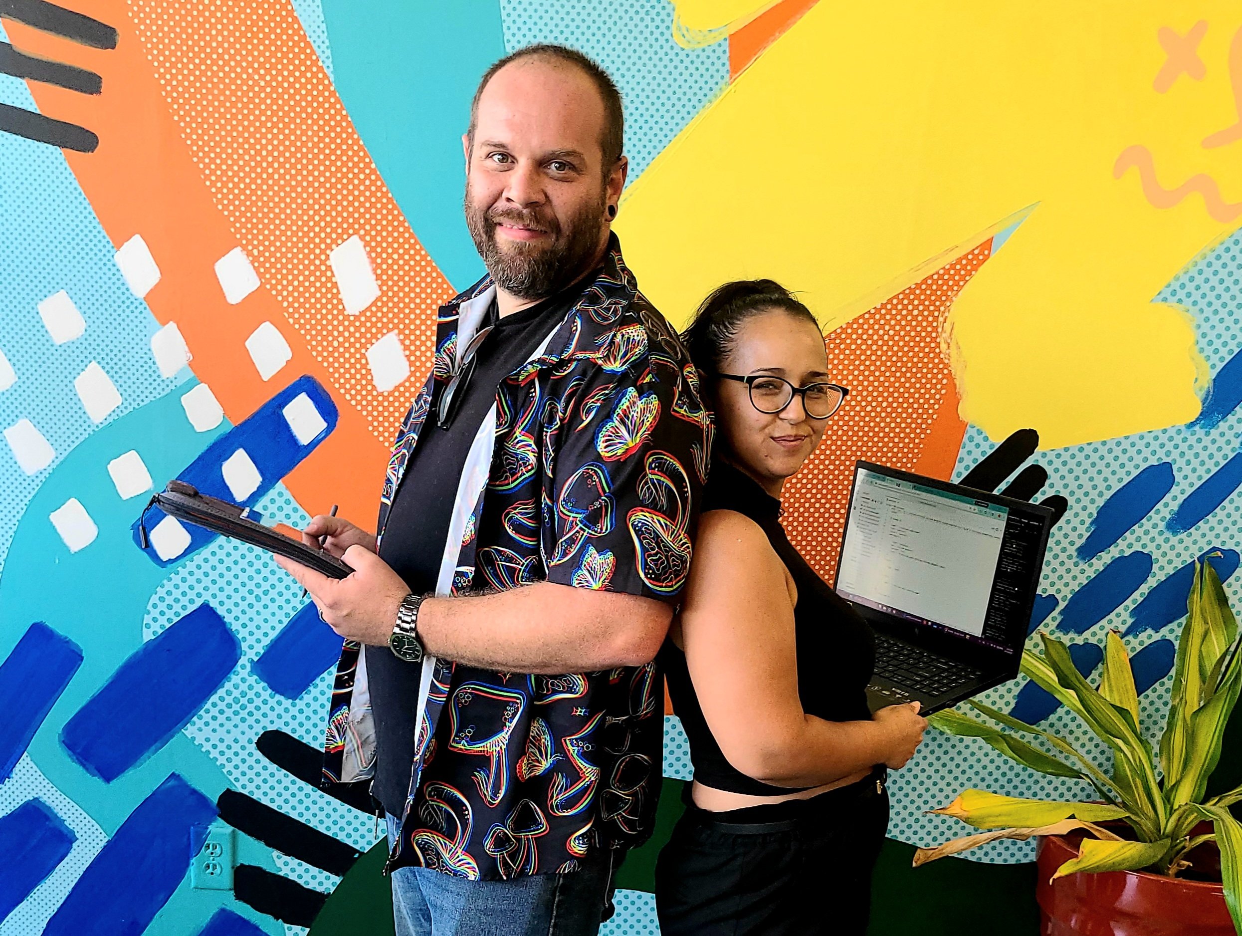 Man and woman holding laptops in front of a colorful mural