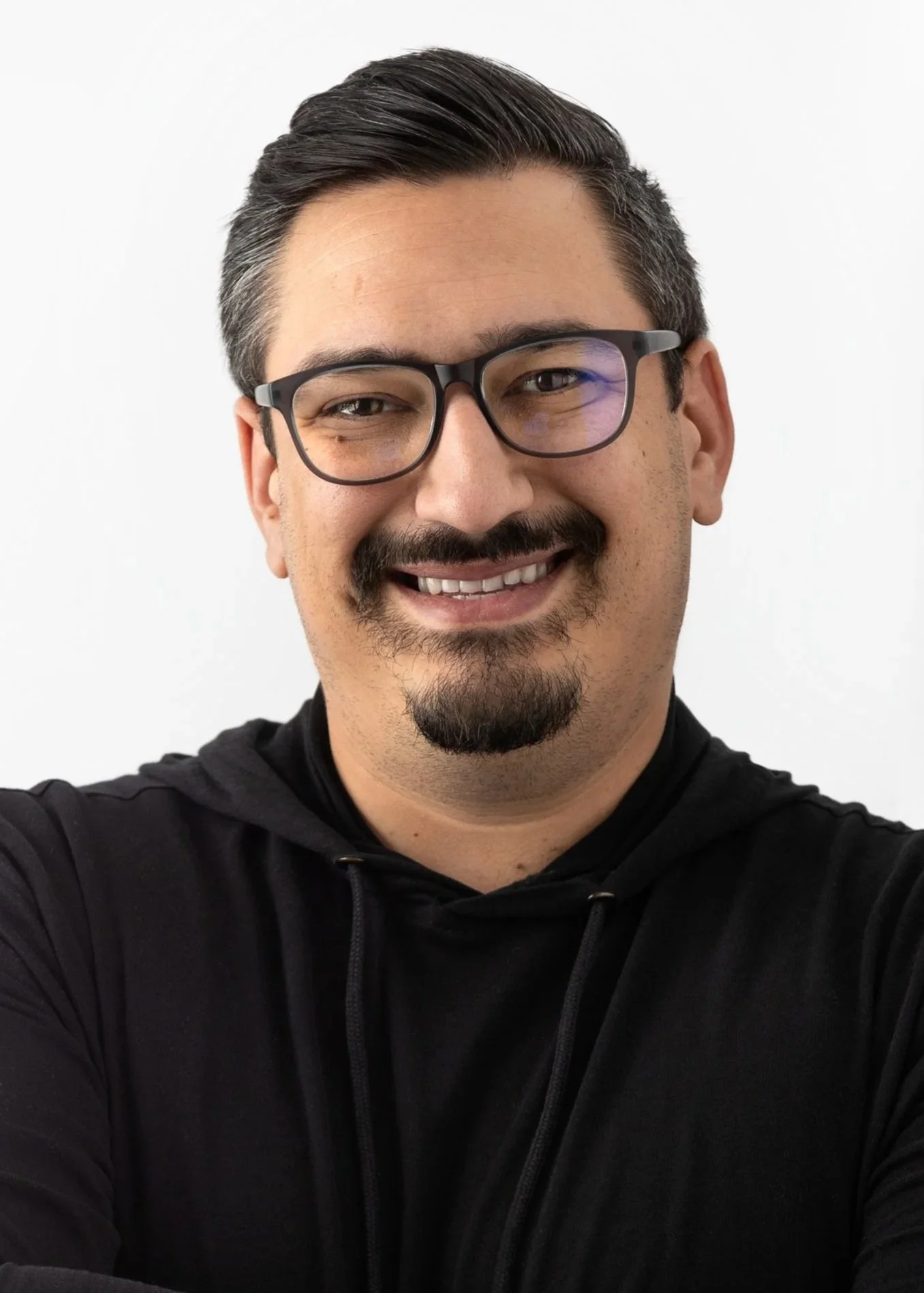 A smiling man with short dark hair, glasses, facial hair, and wearing a black hoodie, taking a selfie against a plain white background.