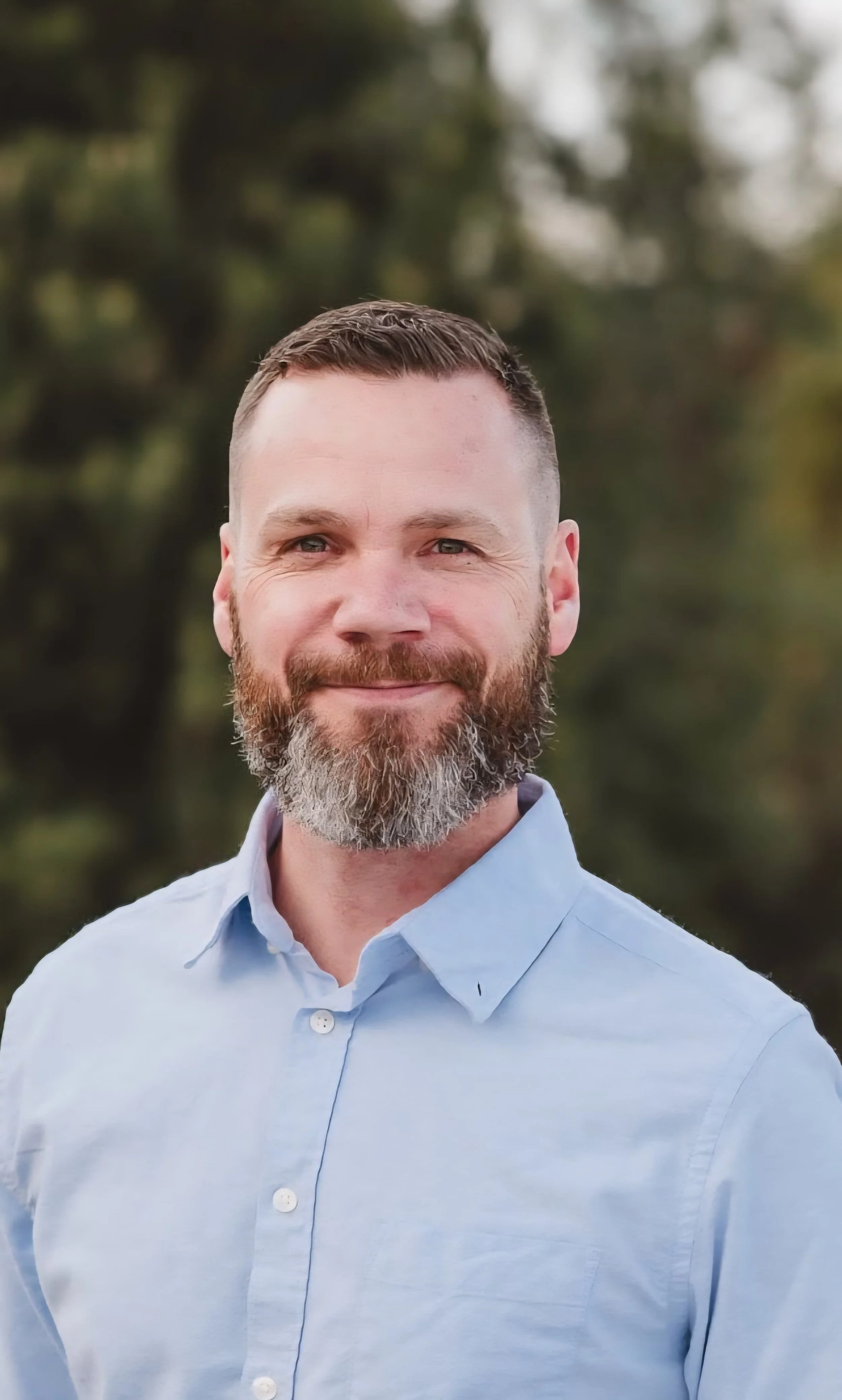 A man with a neatly trimmed beard and short brown hair standing outdoors, wearing a light blue button-up shirt, smiling with a blurred green and brown natural background.