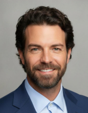 A man with dark, curly hair and a beard, wearing a blue suit and white shirt, smiling at the camera.