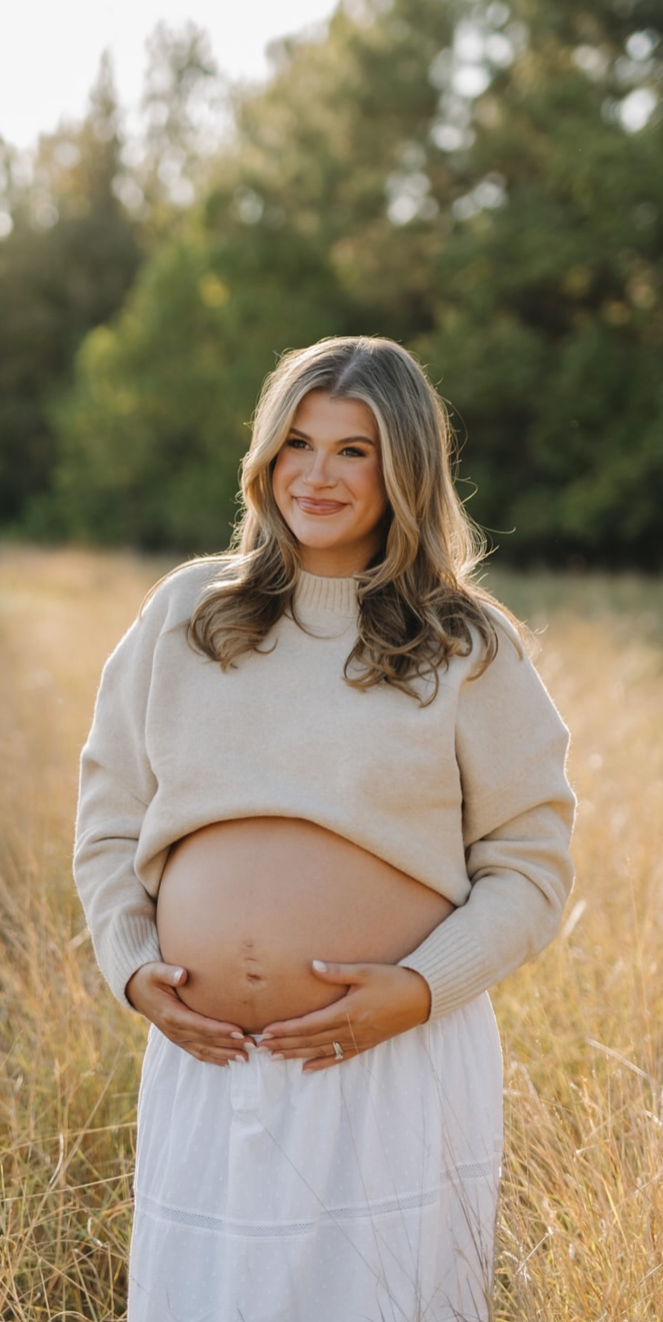 A pregnant woman with long hair, wearing a beige sweater and a white skirt, standing in a field of tall grass with trees in the background, smiling and holding her belly.