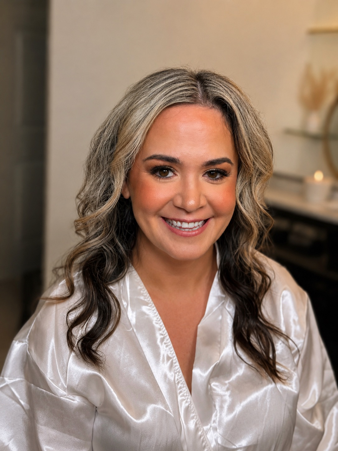 A smiling woman with wavy blonde and brunette hair, wearing a shiny satin white robe.