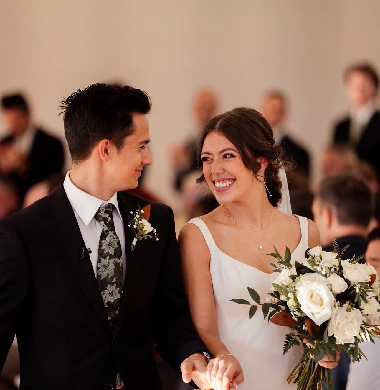A bride and groom smiling at each other during their wedding ceremony, with the bride holding a bouquet of white roses and greenery.