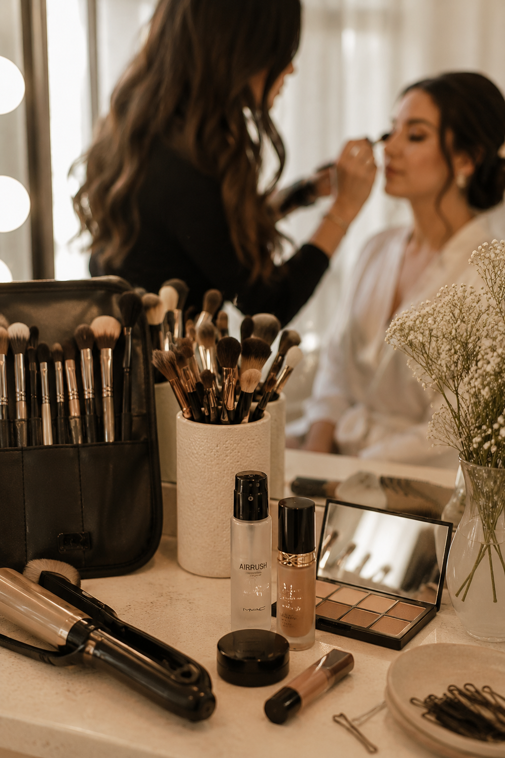 A woman getting her makeup done by another woman in a bright room. On the table in front are makeup brushes, skincare bottles, an eyeshadow palette, and a mirror. The background shows a blurred woman applying makeup to a bride.