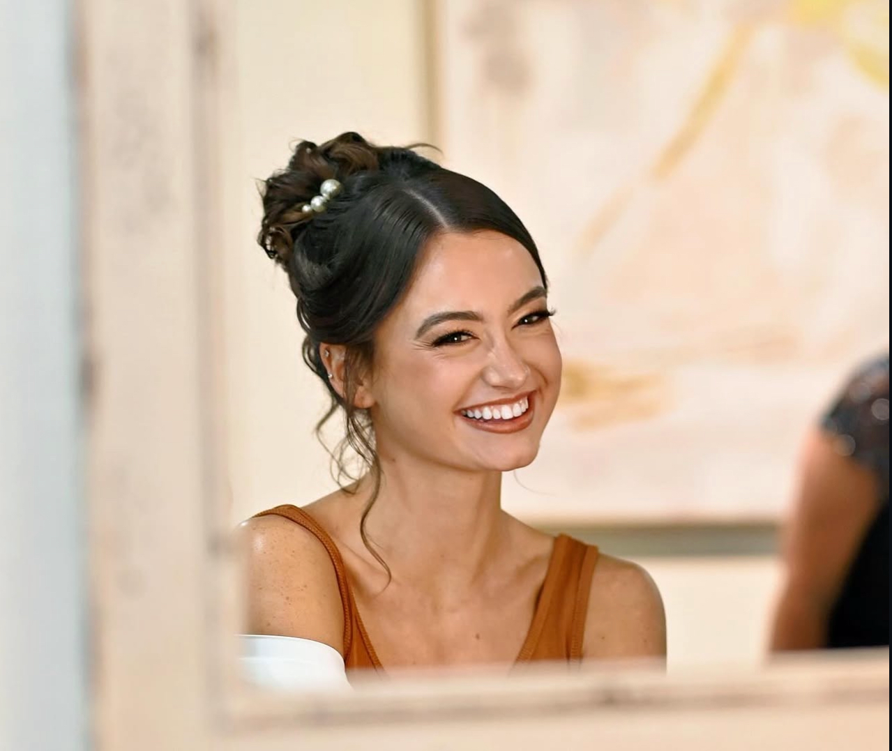 A woman with dark hair styled in an elegant updo with pearl accessories, smiling in a warmly lit room.