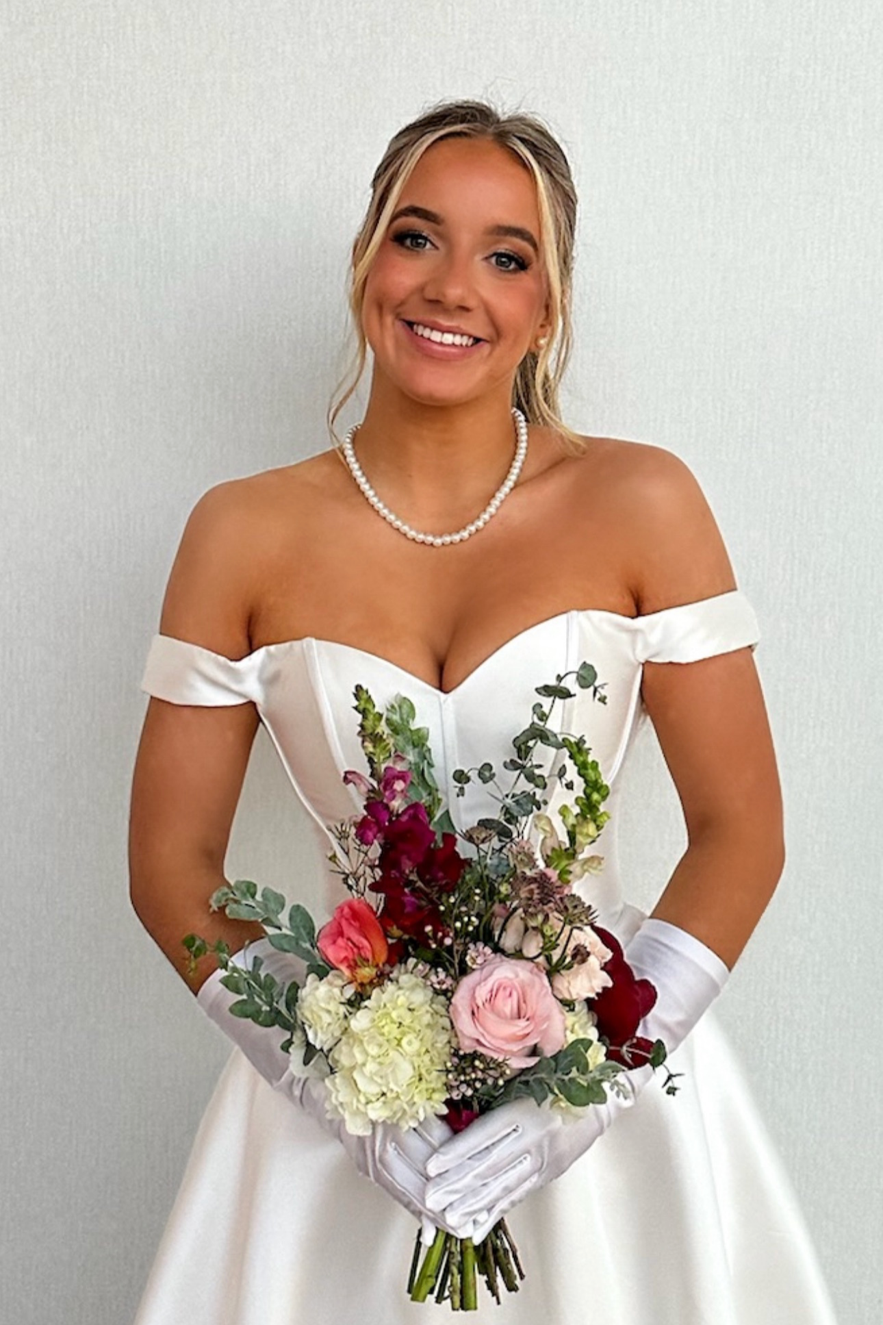 A smiling woman in a white off-shoulder wedding dress holding a bouquet of pink, red, white flowers and greenery against a plain light-colored background.