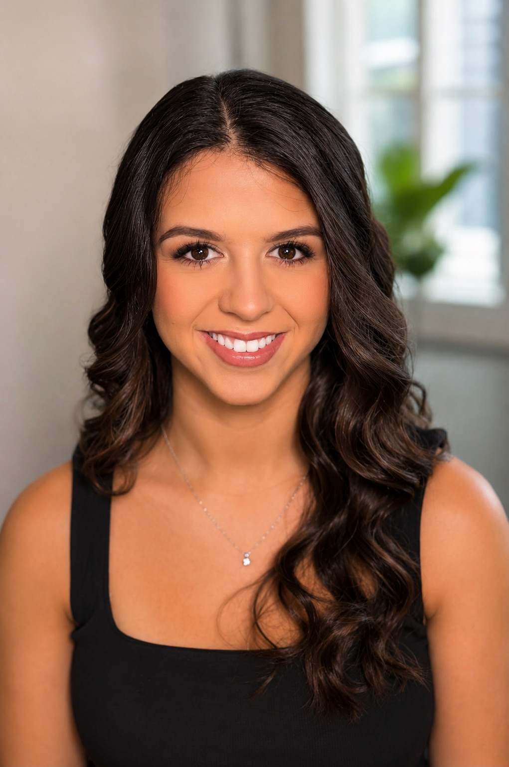 Portrait of a young woman with long wavy dark hair, smiling, wearing a black sleeveless top and a silver necklace, in front of a blurred background with a window and green plant.