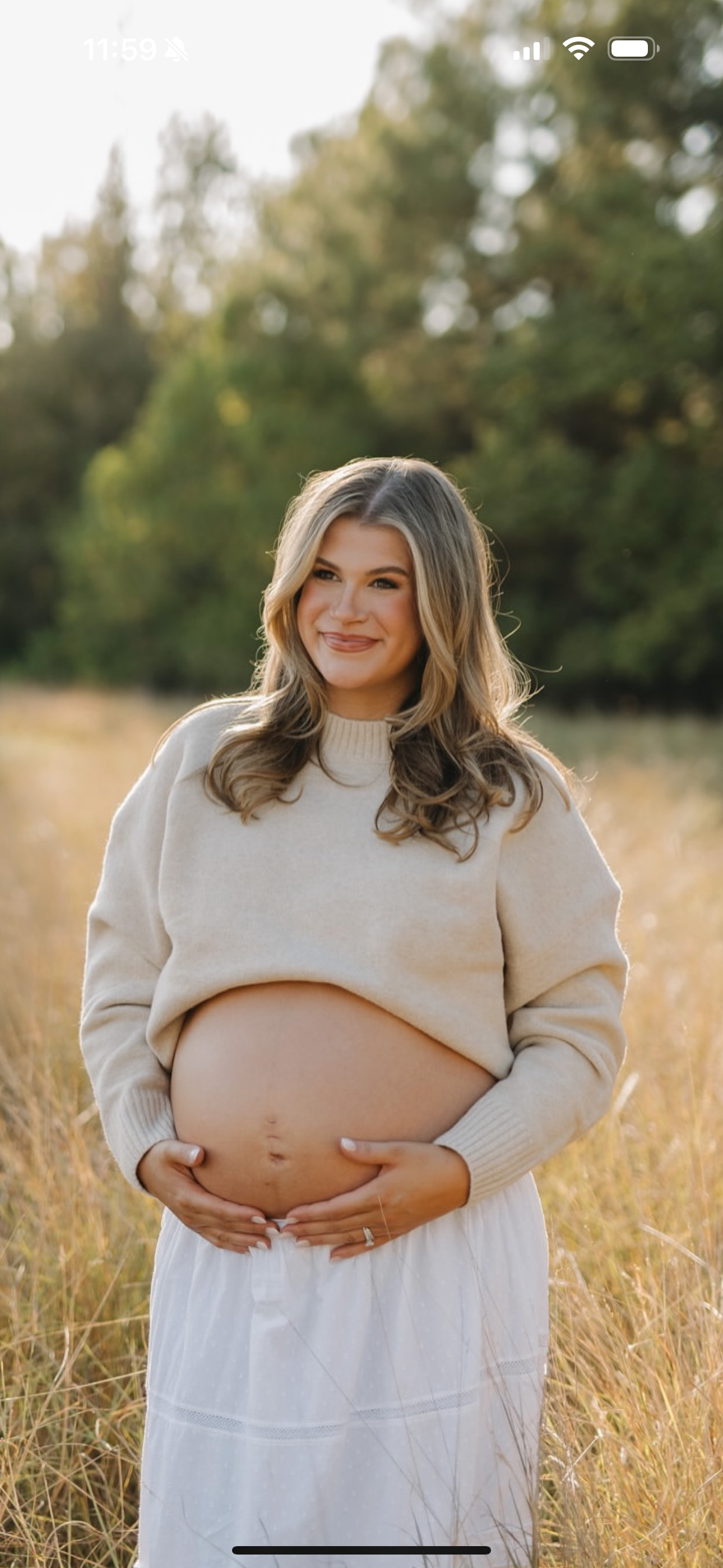 A pregnant woman standing in a field of tall grass during golden hour, smiling and holding her belly with both hands, wearing a beige sweater and a white skirt with a natural background of trees.