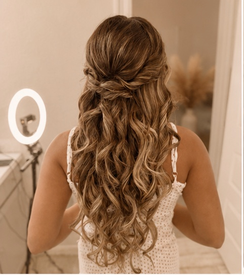 Woman with long, curly, styled hair standing in a room with a ring light and vanity mirror.