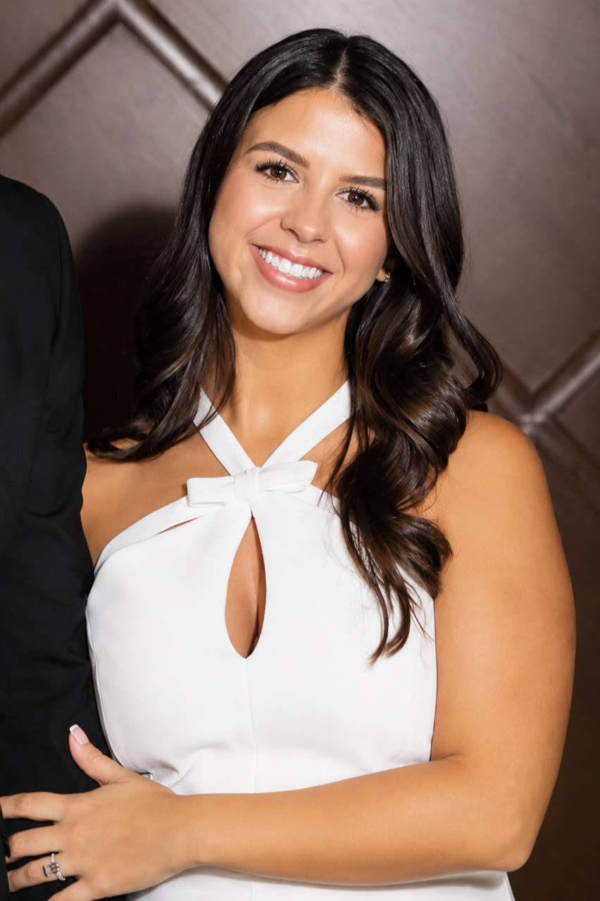 A smiling woman with long, dark hair wearing a white dress with a bow and keyhole neckline, standing against a wooden background.