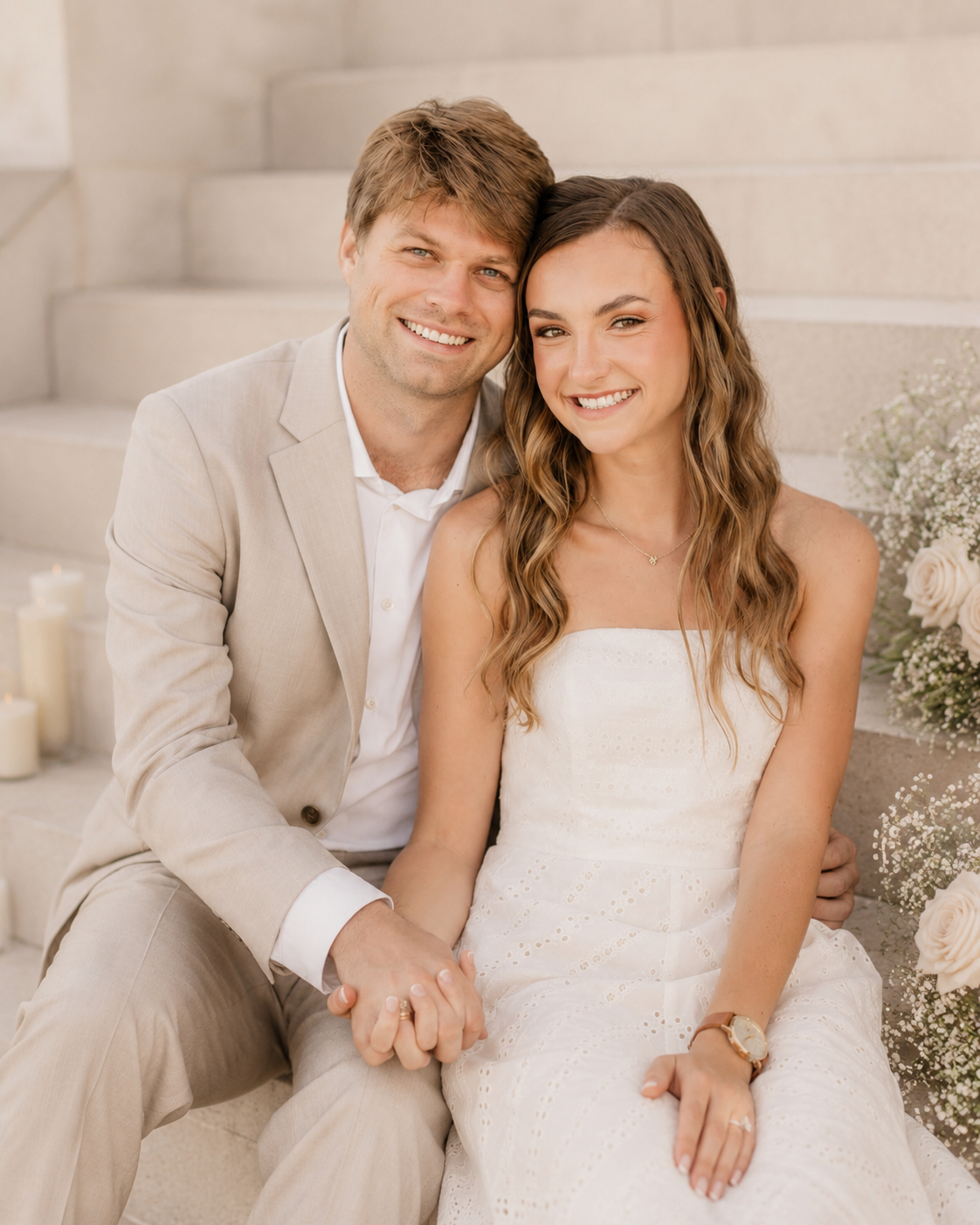 A smiling young woman in a white dress and a young man in a beige suit sitting on steps, holding hands and surrounded by flowers and candles.