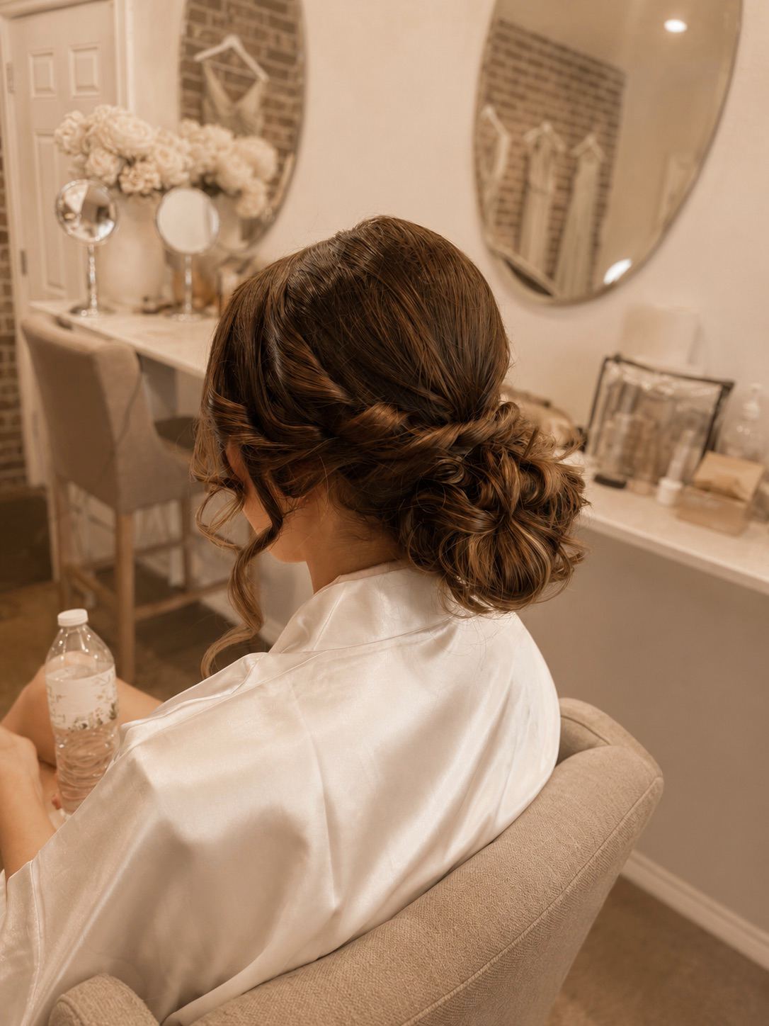 A woman with styled brown hair in an elegant updo, sitting in a beige chair in a beautifully decorated room with floral arrangements and mirrors.