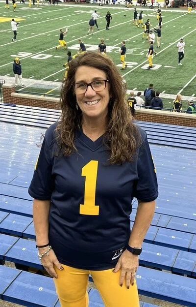 A woman with long curly hair, glasses, and a big smile, wearing a navy blue football jersey with the number 1 and yellow pants, standing in front of a football stadium with players practicing on the field in the background.
