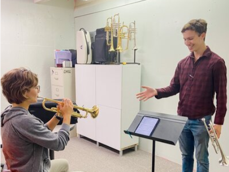 A young boy practicing trumpet lessons with an instructor in a music room, with brass instruments in the background.