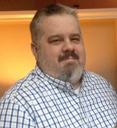 A man with short, light brown hair and a beard, wearing a blue and white checkered shirt, standing indoors with warm lighting.