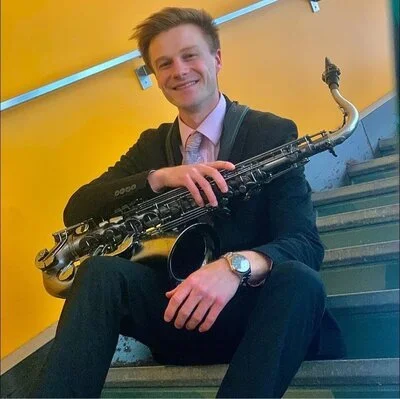 A man in a suit sitting on stairs, holding a saxophone, smiling, with a yellow wall in the background.