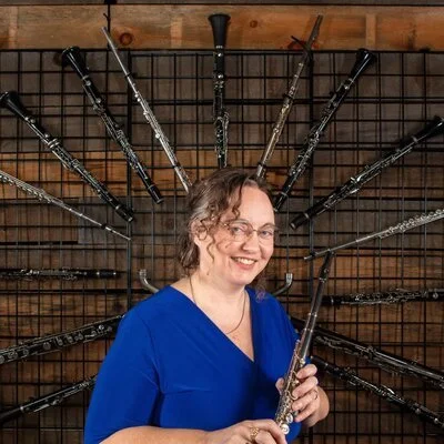 Woman in blue scrubs holding a flute, standing in front of a wall display of various musical instruments.