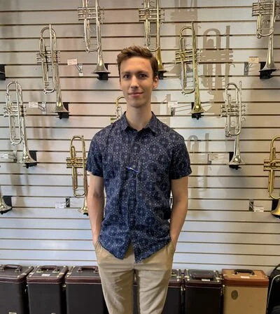 Young man standing in a music store, surrounded by brass and wind instruments such as trumpets and trombones on display.
