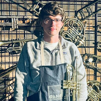 Young man wearing glasses, a gray hoodie, and an apron, standing in front of a wall display of various brass instruments, including trumpets and trombones.