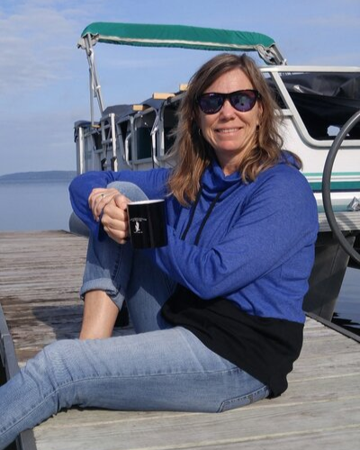 A woman sitting on a wooden dock near a boat, holding a black mug, wearing sunglasses, a blue hoodie, and jeans, with water and a cloudy sky in the background.