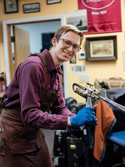 A young man with glasses, smiling, wearing a burgundy shirt, brown apron, and blue gloves, working with equipment in what appears to be a workshop or store.
