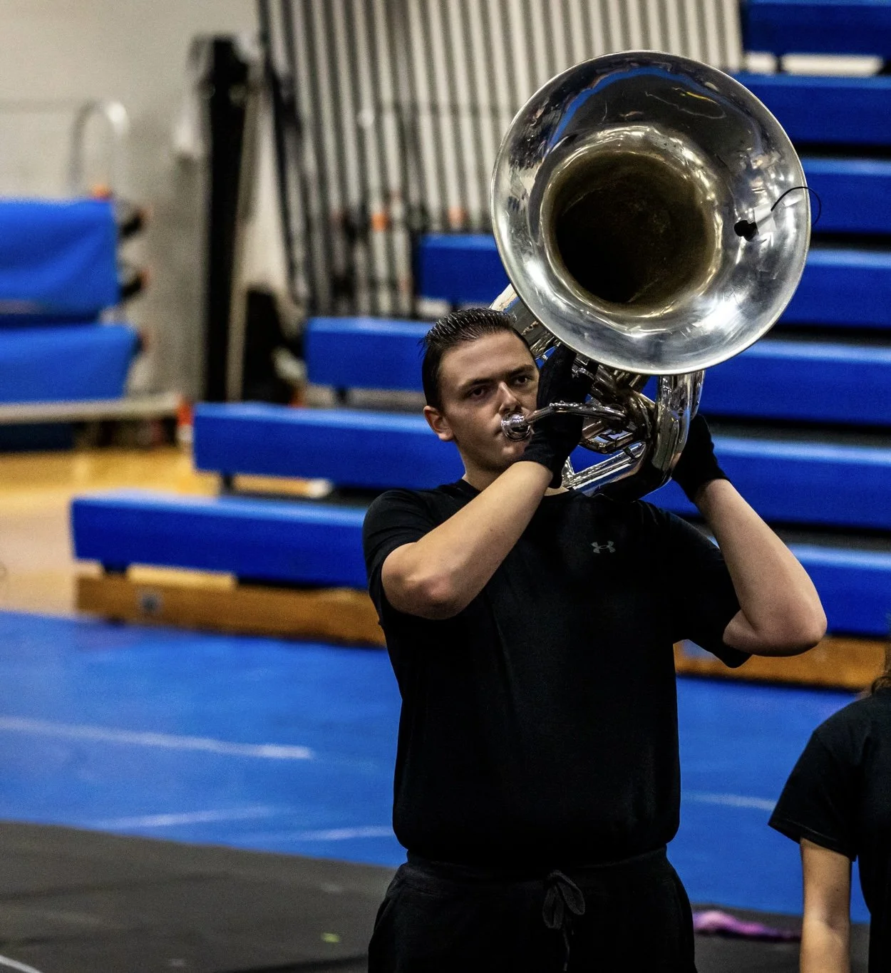 A young man in black shirt and gloves playing a silver sousaphone in a gymnasium with blue padded walls and bleachers in the background.