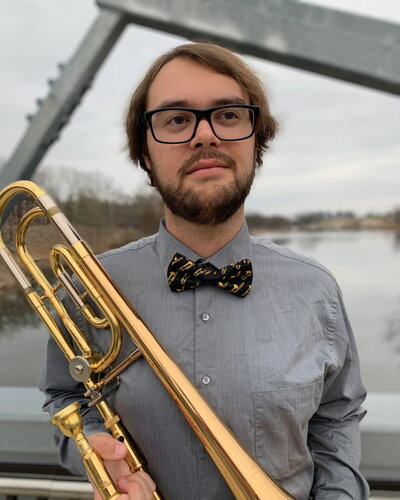 A young man with glasses and a bow tie holding a trombone outdoors by a body of water.