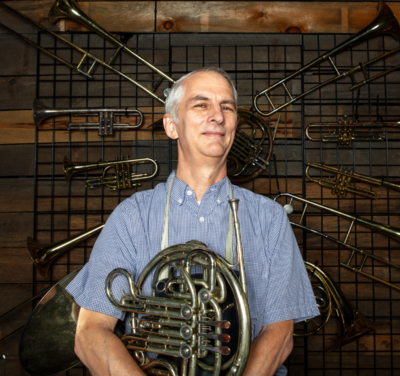 A man holding a French horn, standing in front of a wall with multiple brass instruments hanging on it.