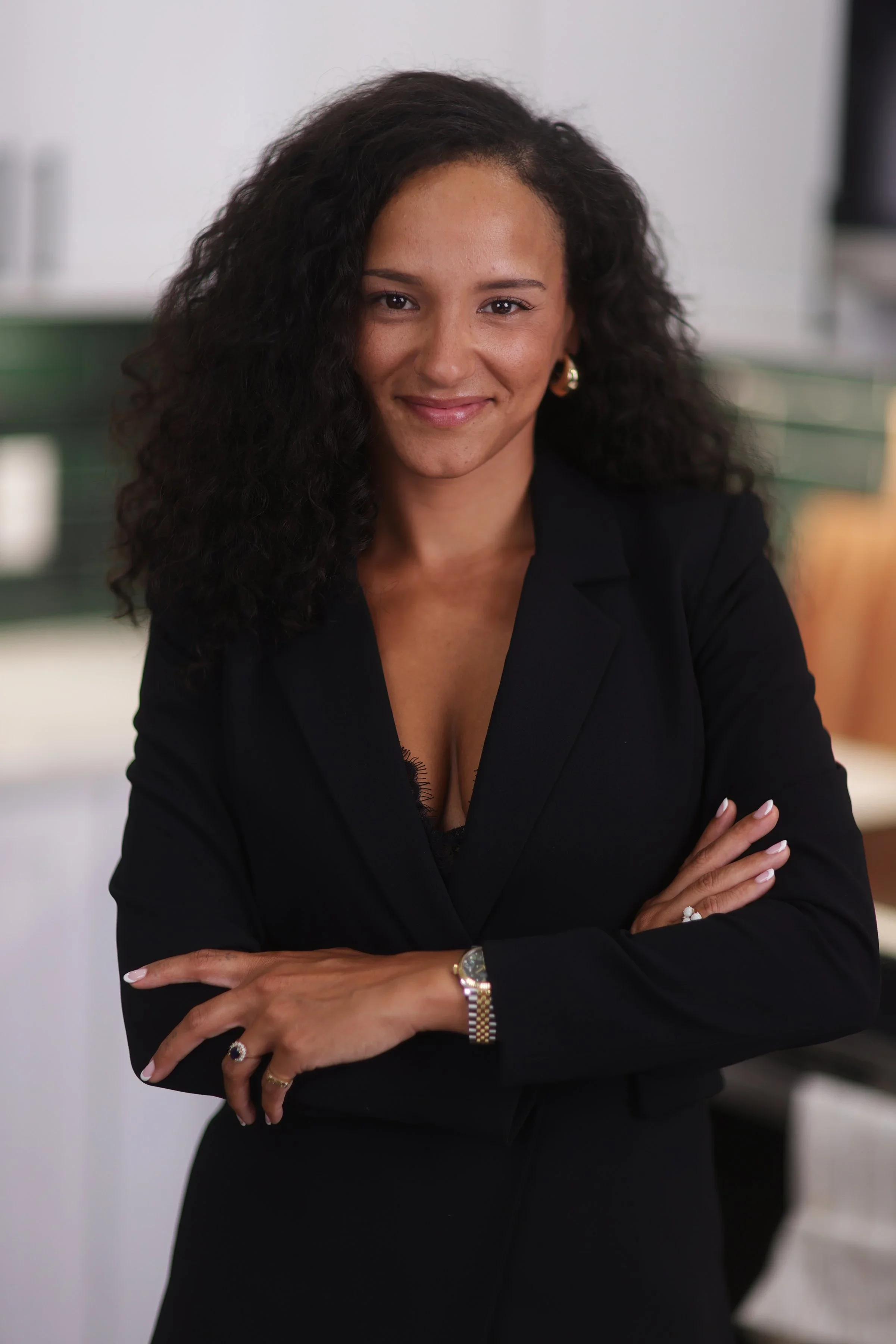 A woman with curly dark hair, wearing a black blazer, smiling with arms crossed in an office setting.