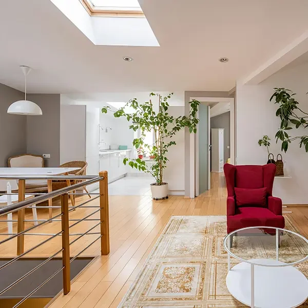 Bright living room with skylight, white walls, wooden flooring, red armchair, potted plants, glass coffee table, and a dining area with a table and chairs.