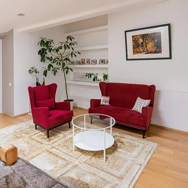 Living room with red upholstered armchair and sofa, white round coffee table, bookshelf with framed photos, large potted plant, framed landscape picture on white wall, and wooden floor rugs.
