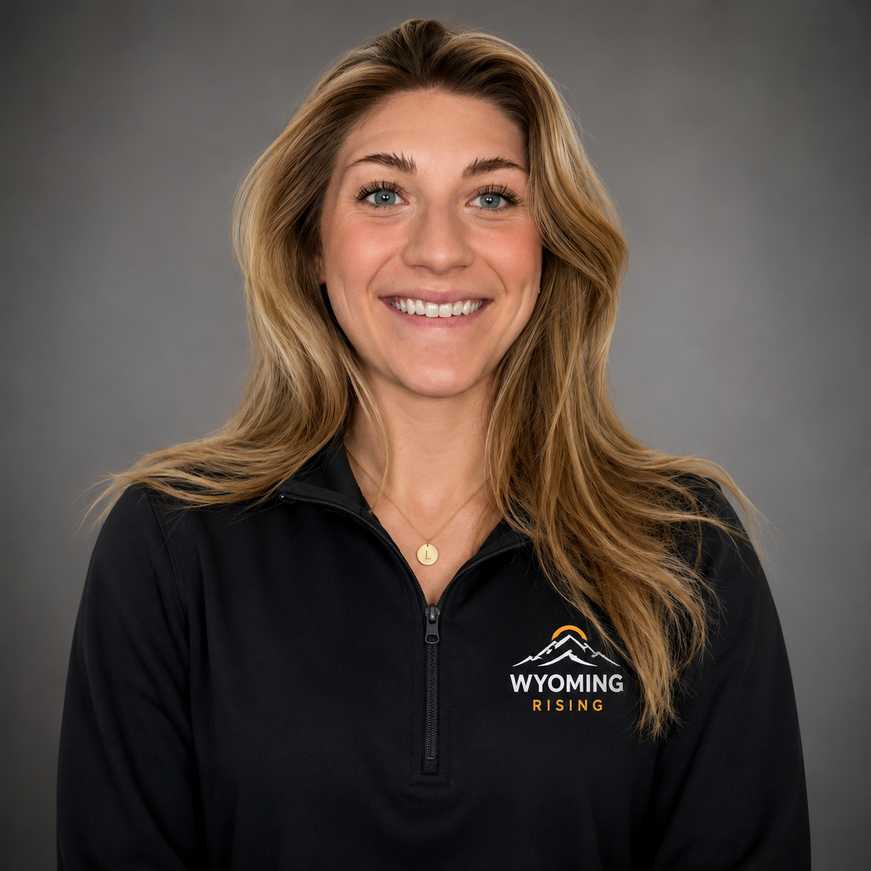 A smiling woman with long, wavy blonde hair, wearing a black Wyoming Rising jacket and a gold necklace with a circular pendant.