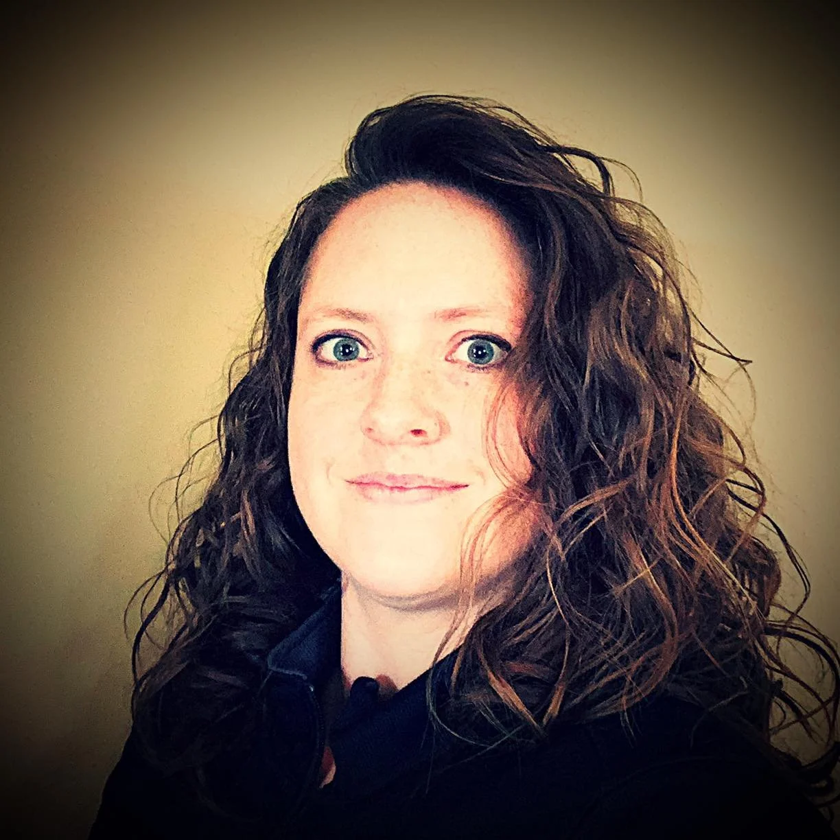 A woman with curly brown hair, blue eyes, and light skin, smiling slightly, wearing a black top, standing against a plain beige background.
