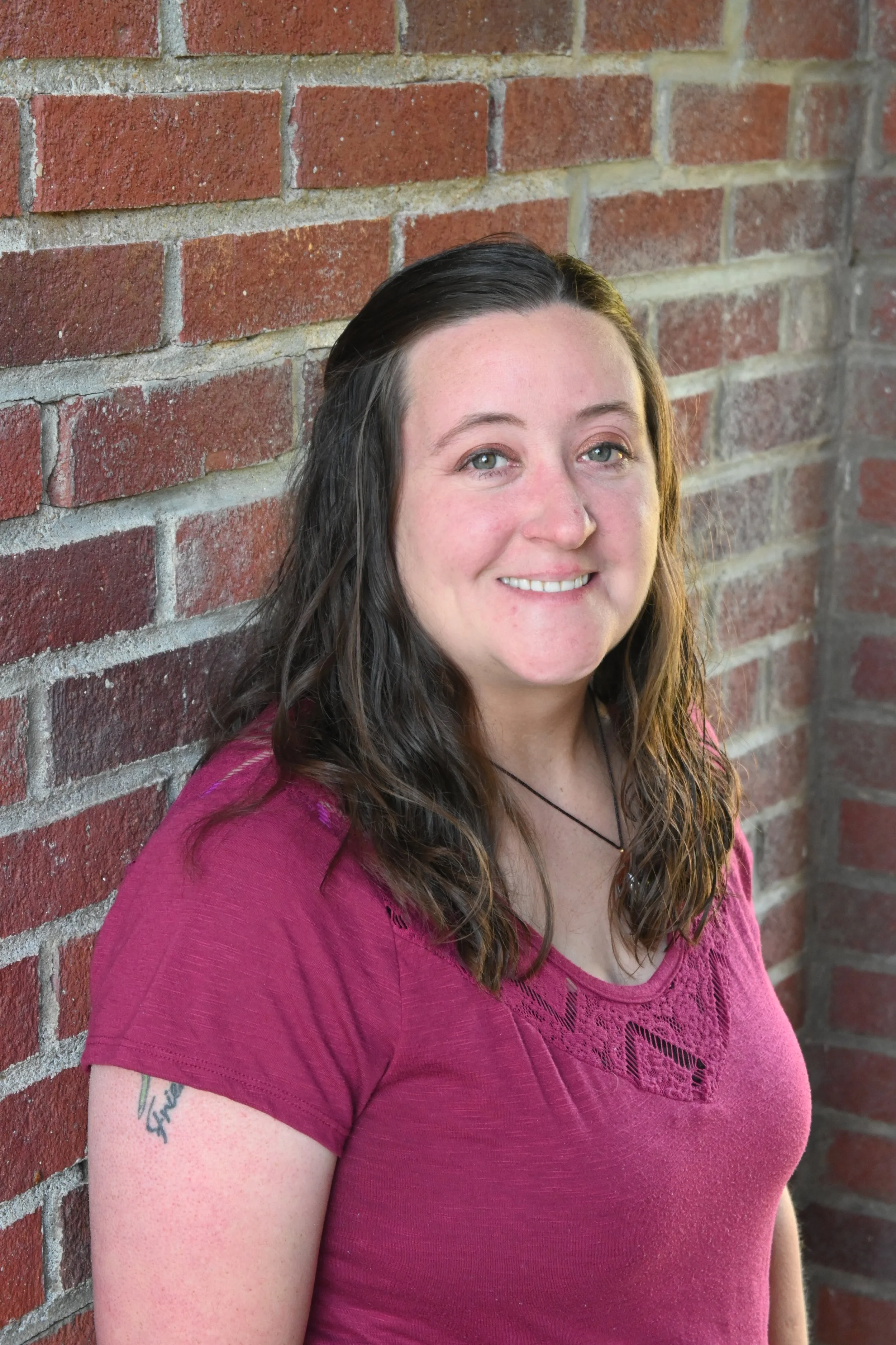 A woman with long, wavy brown hair and light skin is smiling, standing against a red brick wall, wearing a pink shirt.