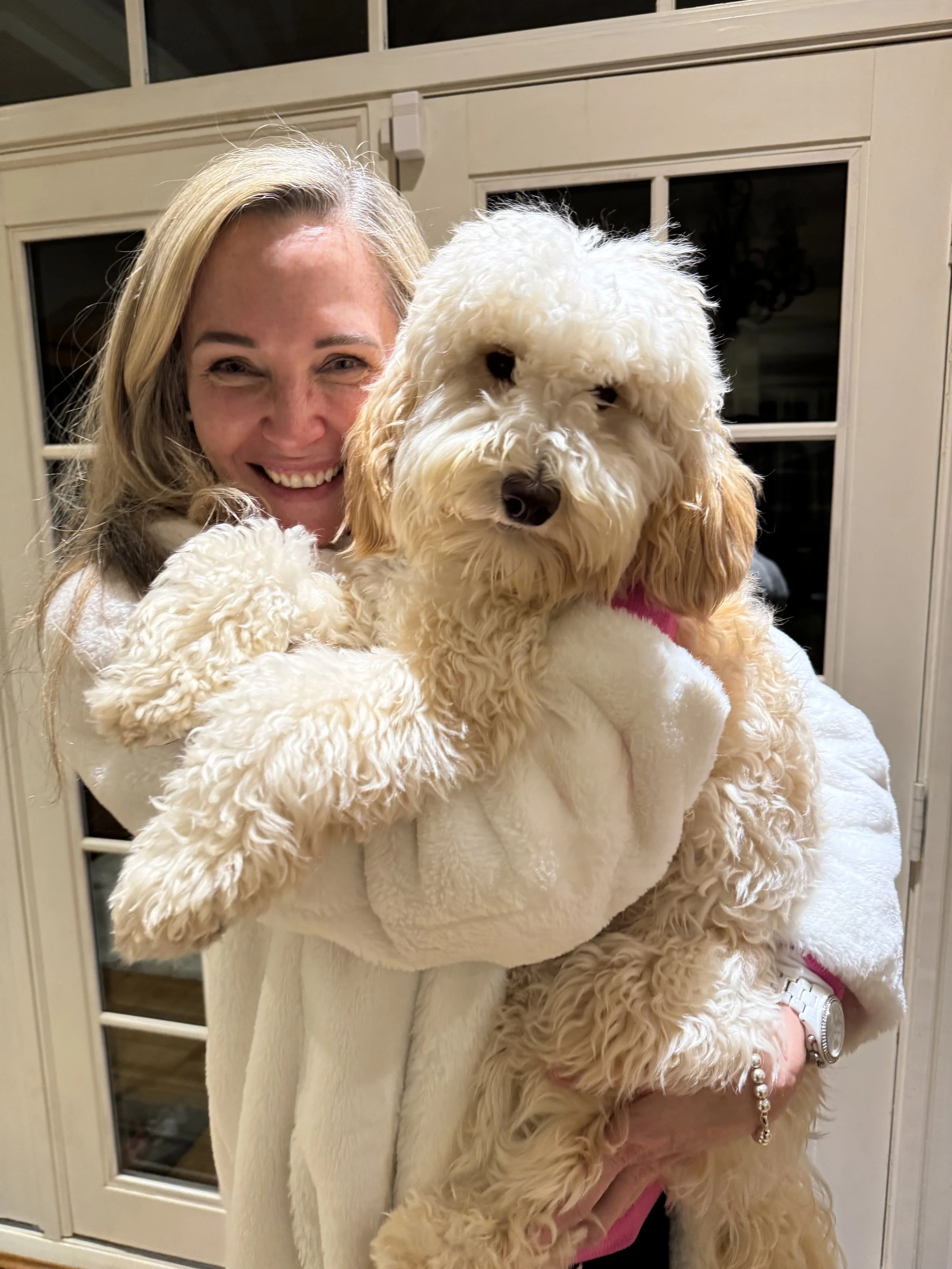 A woman with blonde hair smiling while holding a large, fluffy, cream-colored dog in her arms inside a house near a glass door.