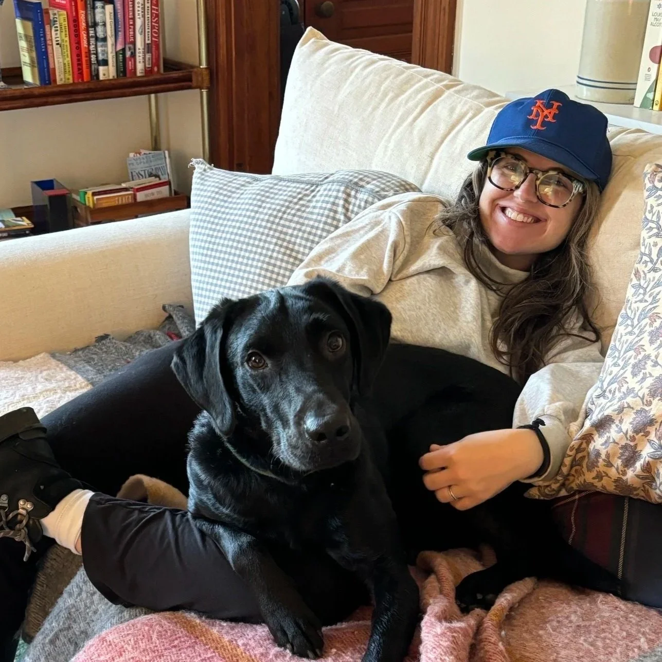 A woman with long brown hair, glasses, and a blue New York Mets cap is lying on a couch with a black Labrador Retriever puppy, smiling at the camera.