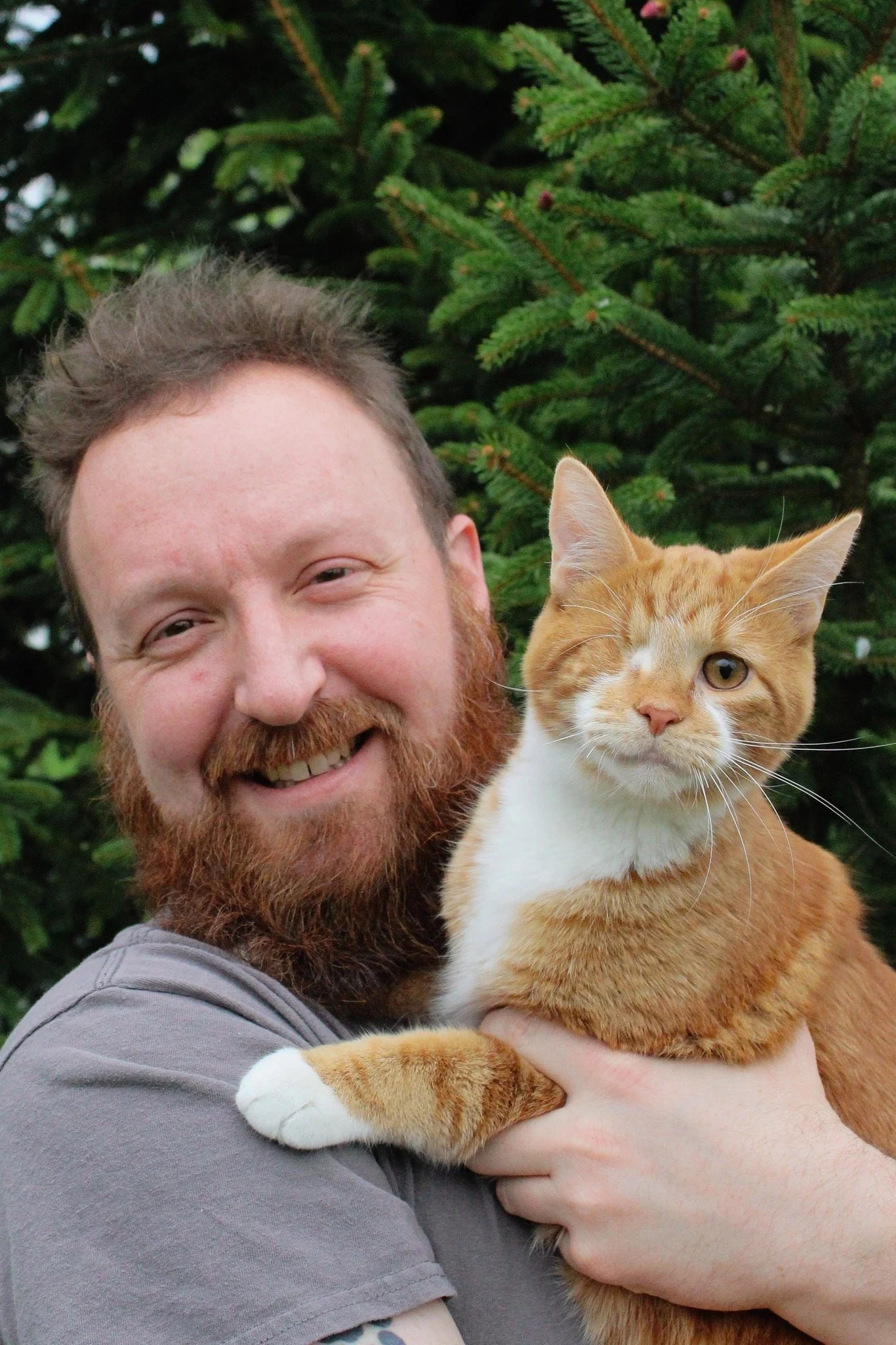 A smiling man with a beard holding an orange and white cat, standing in front of a green pine tree.