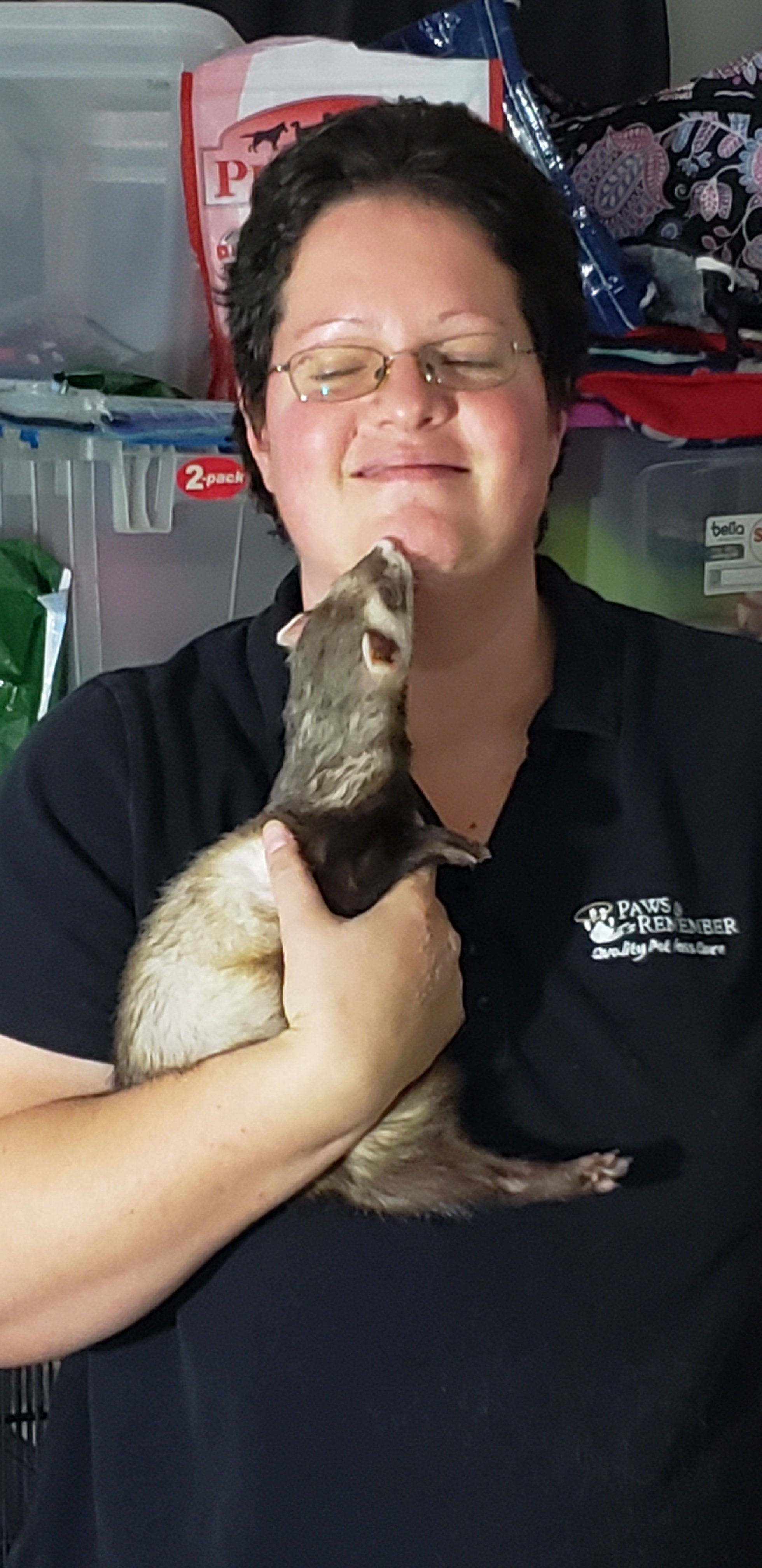 A woman holding a ferret close to her face, smiling with her eyes closed, in a room with pet supplies in the background.