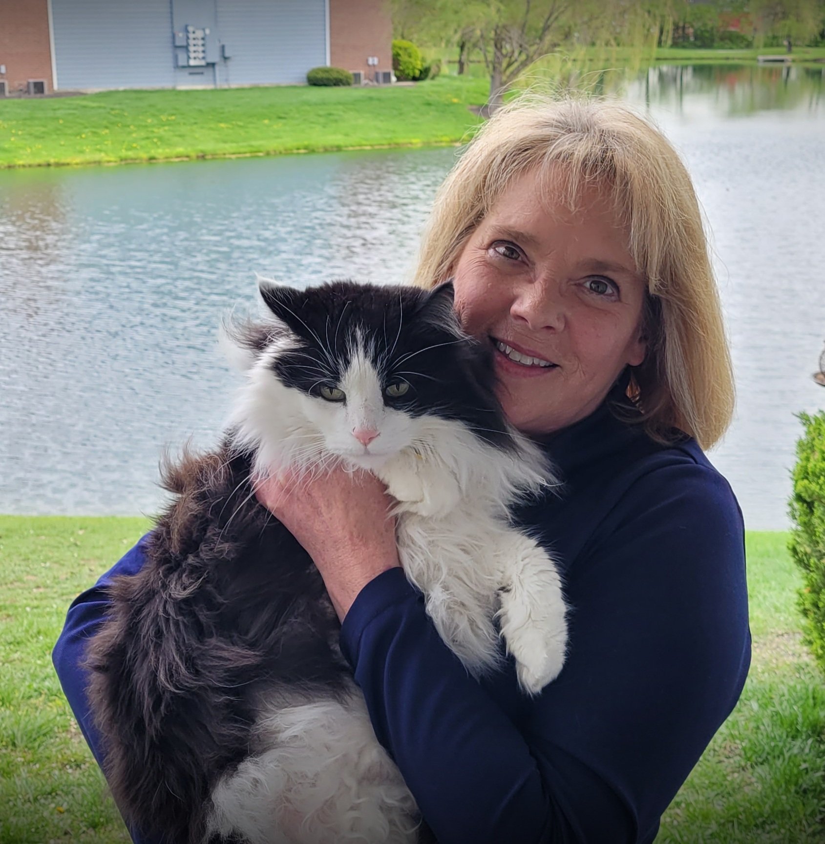 Woman holding a black and white cat by a lake with green grass and trees in the background.