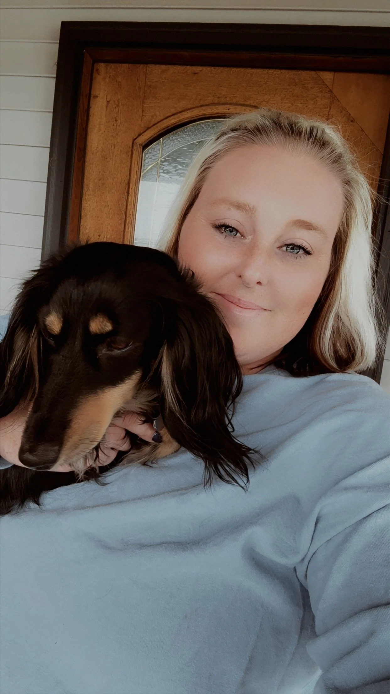 A woman with blonde hair and blue eyes hugging a black and tan long-haired dachshund in front of a wooden door.
