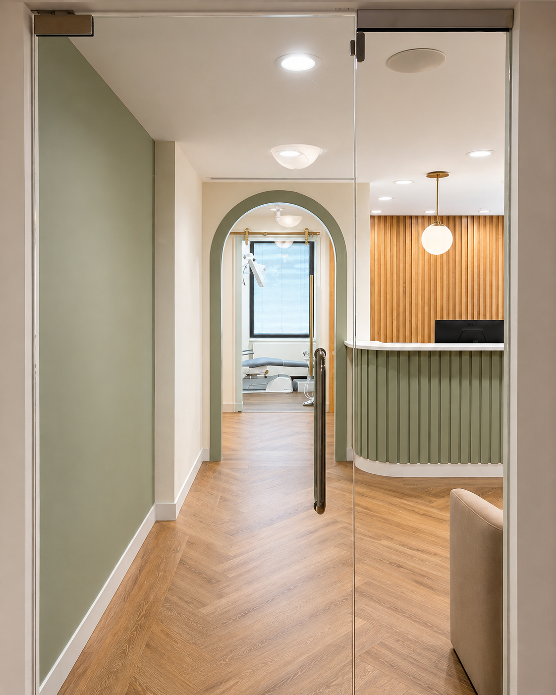 Dental reception area with green and wooden accents, glass door, and hallway leading to a room with medical equipment and a treatment table.