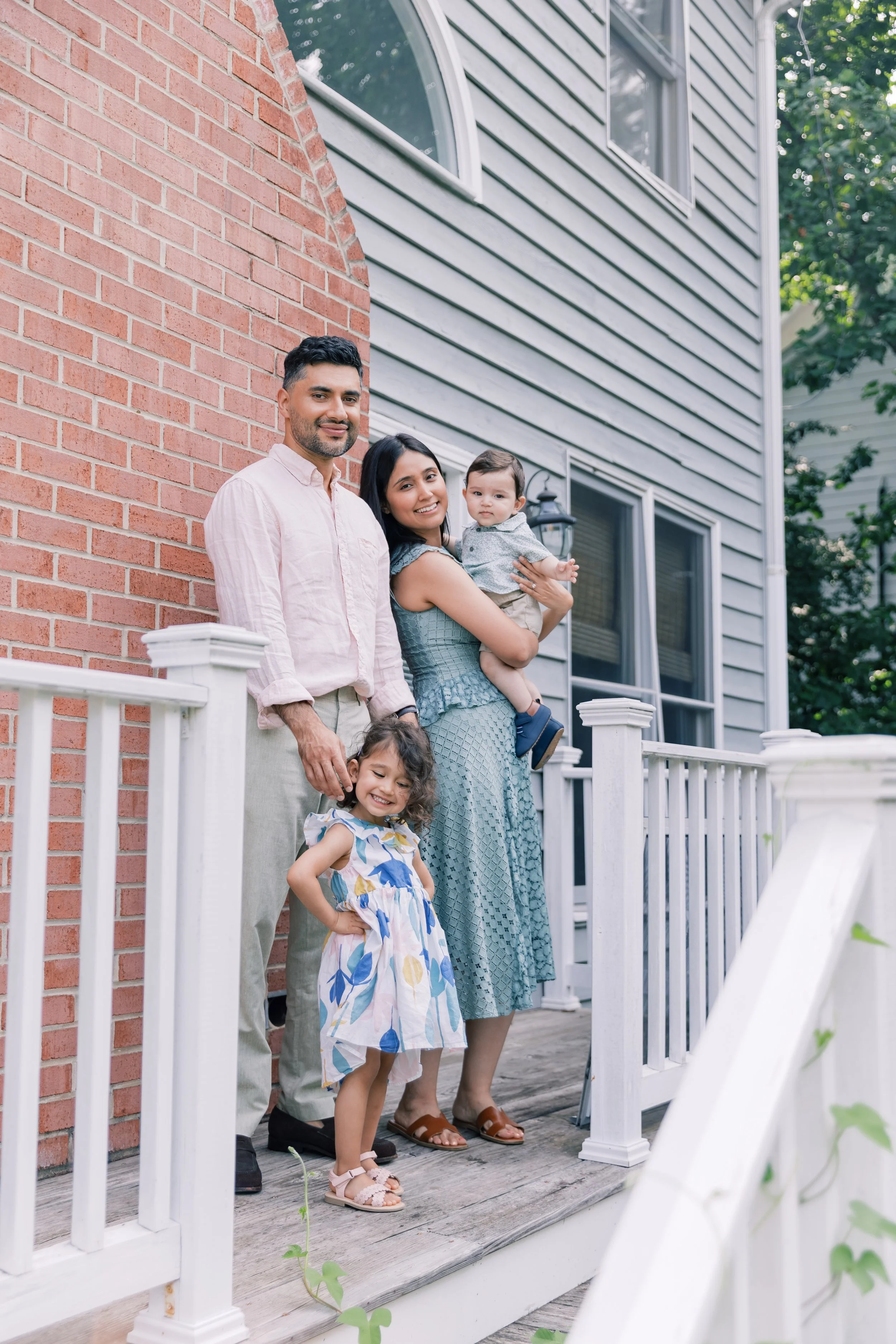 A family of five stands on a front porch with white railing and a brick and siding house behind them. The dad is on the left, the mom holding a young boy, and one girl is in front.