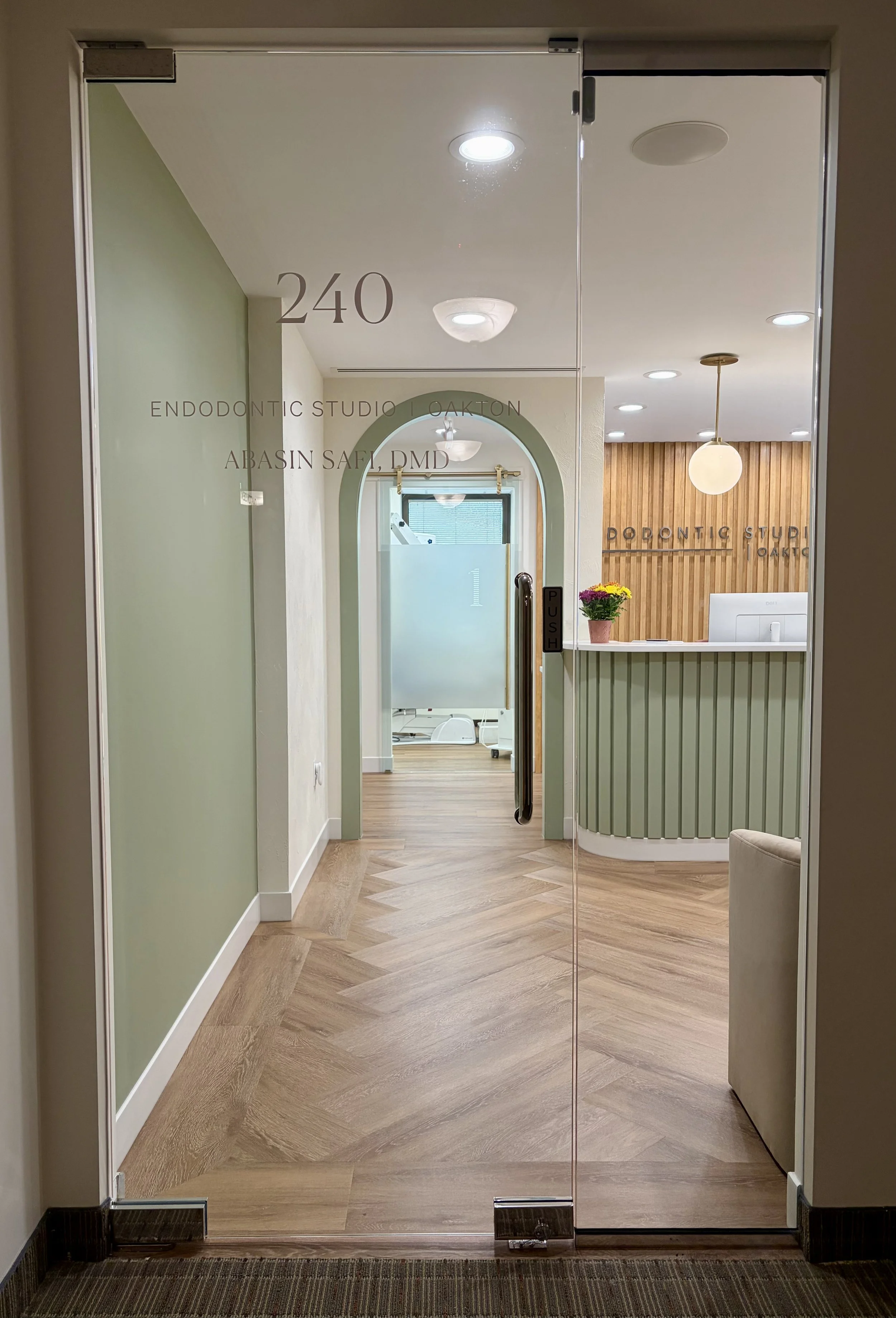 Dental reception area with green and wooden accents, glass door, and hallway leading to a room with medical equipment and a treatment table.